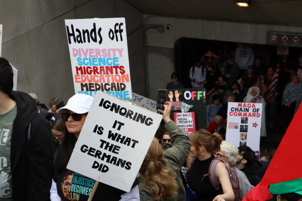 Demonstrators hold up multicolored signs while standing on an escalator leading out of the Smithsonian metro station. One sign writes "Ignoring it is what the Germans did," another sign writes "Hands off diversity science migrants education medicaid palestine," another sign in black writes "oligarchs vs the rest of us," and another sign writes "putin's wish list economy wrecked alliances destroyed populations unvaxxed" with checkmarks to the left of the three listed items.