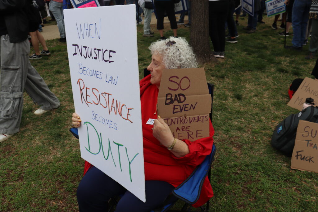 A demonstrator dressed in red sits on a lawn chair and holds up two signs. The larger sign is white and to the left of the picture. It writes "when injustice becomes law resistance becomes duty." The sign to the right of the picture is cardboard and writes "so bad even introverts are here."