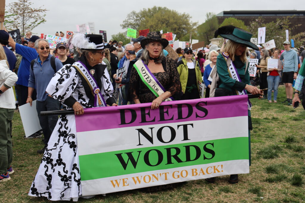 Three demonstrators dressed in suffragette costumes hold up a banner. The banner is purple white and green. On the banner, there are the words "deeds not words" and the yellow words "we won't go back!"